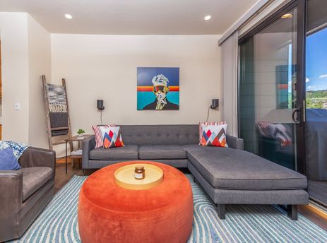 The living room at Basecamp Luxe features a gray sectional sofa with colorful throw pillows, an orange ottoman, and a modern art piece on the wall.