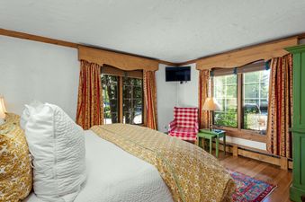 The bedroom at Chateau Christian 2 Bedroom Gold Unit 110 boasts a comfortable bed with white and gold bedding, complemented by red and gold patterned curtains, a checkered red and white armchair, a wall-mounted TV, and a green side table with a lamp.