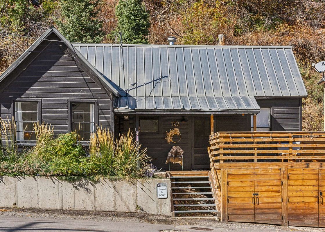 The exterior of Daly Cottage, a charming dark-sided home with a metal roof, front entrance steps, and a wooden fence, nestled into a hillside landscape.