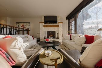 A cozy living room at Ivory Stone Chalet at See Forever with a roaring fireplace, plush white sofas with red and white pillows, and a large window offering a view of a winter landscape.