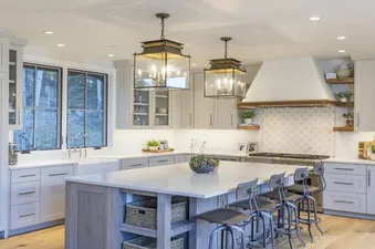A modern kitchen with light gray cabinetry, white countertops, a large island with seating, and two large pendant lights, with an outdoor view through windows.