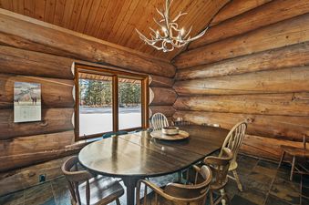 A rustic dining area at the Stillwater Log Home with a large oval wooden table and antler chandelier, featuring log walls and a window overlooking a snowy, tree-filled landscape.