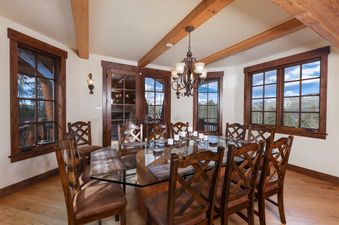 The Fox Paw Lodge features an exquisite dining area with a glass-top table, elegant wooden chairs, and large windows offering panoramic views of the mountains and lush trees.