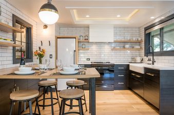 A kitchen with black cabinets, white subway tile, and a farmhouse sink. There is a small breakfast bar with stools.