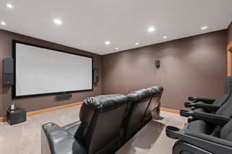 A home cinema room with a large projection screen, two rows of black recliners, and surround sound speakers, set against warm brown walls and a carpeted floor.