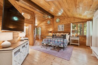 A cozy king bedroom at Apres Chalet, featuring wood-paneled walls and ceiling, a dark metal bed frame with a colorful blanket, two striped ottomans, a wall-mounted TV over a distressed white cabinet, and side tables with lamps, all bathed in soft light fr