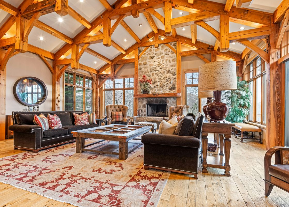 A spacious living room at Oak Wood Lodge with a vaulted wooden beam ceiling, stone fireplace, comfortable seating, and large windows.