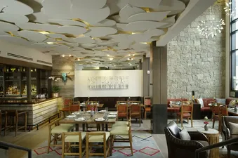 A stylish restaurant interior featuring dining tables, a fully stocked bar, and unique ceiling decor with leaf-like panels, bathed in natural light.