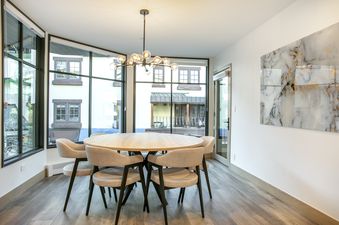 The dining area within the Vail Center Place 2 Bedroom Diamond Unit 2 features a round wooden table, surrounded by six beige chairs, beneath a stylish chandelier with multiple globes, and large windows offering views of the exterior buildings.