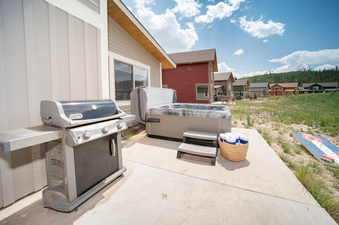 The patio at Alder Brook Homers Hideaway features a hot tub, BBQ grill, and a basket of towels, set against a backdrop of distant homes and lush green hills.
