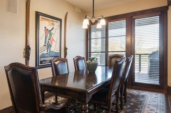 A rustic dining area with a large wooden table, surrounded by leather chairs, under a hanging chandelier, with a ski-themed poster on the wall.