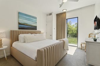 A modern bedroom at The Mason 1052 features a beige upholstered bed with white bedding, an abstract painting, and a private patio door leading to a green lawn.