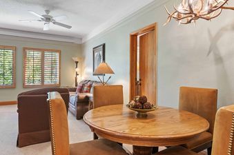 A cozy dining area with a round wooden table and seating connects to a living space with comfortable sofas, featuring a ceiling fan and natural light from shuttered windows.