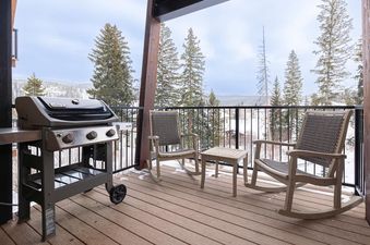A private balcony at the Bullseye Retreat features a BBQ grill, two rocking chairs, and a small table, all set against a winter backdrop of snow-covered evergreen trees and distant mountains.