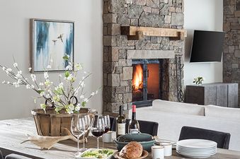 The dining area at Four Pines 06, featuring a rustic wooden table set with food and wine, illuminated by a large circular chandelier, with a stone fireplace and living area in the background.