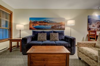 The living area of the Blackcomb Springs Premium Studio features a comfortable blue sofa, a rustic wooden coffee table, and large framed artwork depicting a mountain lake scene, with two lamps providing ambient lighting.
