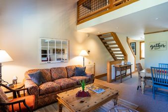 The Northstar Gold Bend Getaway's living room is furnished with a patterned sofa, a wooden coffee table, and an accent mirror, while the adjacent dining area features a wooden table with blue chairs and a 'gather' sign.