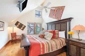 A primary bedroom at Pine Meadows 138 features a dark wood bed with orange and striped bedding, a nightstand with a lamp and flowers, an armchair, and a ceiling-mounted TV, all under a vaulted ceiling with windows offering an exterior view.