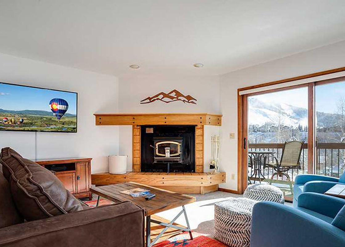 The living room at Ranch at Steamboat RA507 features a brown leather sofa, two blue armchairs, a wooden coffee table, a corner fireplace, and a flat-screen TV, with a sliding glass door leading to a balcony with mountain views.