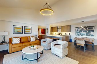 A spacious and modern open-concept living area in Torian Plum Plaza 804, featuring a tan leather sofa, two patterned armchairs, a coffee table, a full kitchen, and a dining area.
