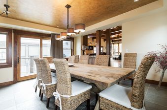 A rustic dining table surrounded by wicker chairs with white cushions is illuminated by a modern linear chandelier in a dining room with warm-toned ceilings and large windows.
