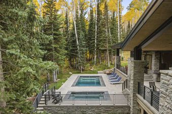 An elevated view of a luxurious outdoor pool and hot tub area surrounded by dense forest with pine and yellow aspen trees, adjacent to a stone and wood modern house.