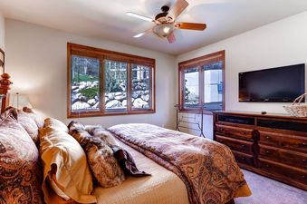 A cozy bedroom at Christie Club 316 with a plush bed, wood dresser, wall-mounted TV, and large windows.