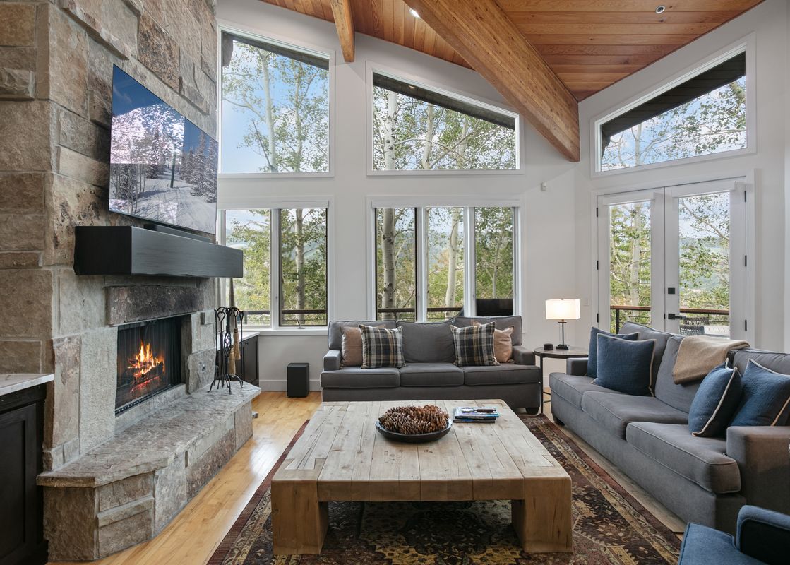 The cozy living room of Twin Pines 5 featuring a large stone fireplace, grey sofas, and beautiful wooded views through tall windows.