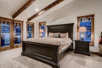 A master bedroom at the Lodge at Ski Hill featuring a large dark wooden bed, two windows with wooden blinds and frames, two nightstands with lamps, and a carpeted floor.