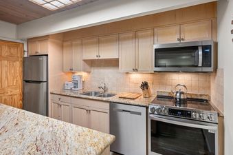 A modern kitchen with light wood cabinets, granite countertops, and stainless steel appliances including a refrigerator, microwave, and oven.