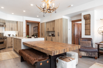 The dining area of 132 Two Bedroom Black Diamond Lodge has a large wooden table with two benches and a cowhide ottoman. The kitchen and living area are visible in the background.