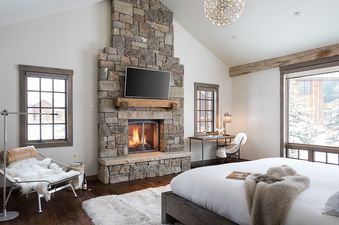 The luxurious bedroom in Four Pines 06 features a stone fireplace with a mounted TV, a comfortable bed, and a desk with a view of a snowy landscape.