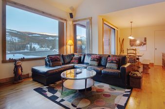 The Snowblaze A33 living room features a large L-shaped leather sofa, a round glass coffee table, and an area rug with a geometric pattern, all set against large windows with a snowy mountain view.