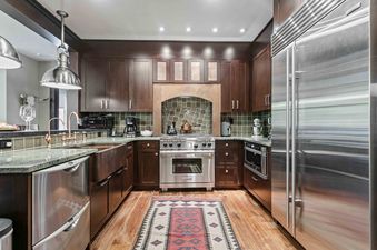 A modern rustic kitchen at Flagstaff 204 featuring dark wood cabinets, green granite countertops, a copper sink, stainless steel appliances, and a red patterned rug on hardwood floors.