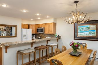 The open concept kitchen and dining area in Montaneros 308 features natural wood cabinetry, granite countertops, a breakfast bar, a rustic dining table with chairs, an antler chandelier, and a framed Vail ski map.