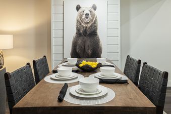 A dining table set with white dishes, black napkins, and placemats, with a bowl of lemons in the center, and a large bear portrait on the wall.