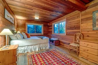 A cozy bedroom with wooden walls and ceiling, featuring a bed with white linens, two windows overlooking a green landscape, a patterned rug, and wooden furniture.