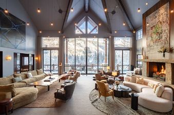 A luxurious living area at The Riverbend House features vaulted ceilings, multiple seating arrangements, a stone fireplace, and large windows with stunning views of snow-covered mountains.