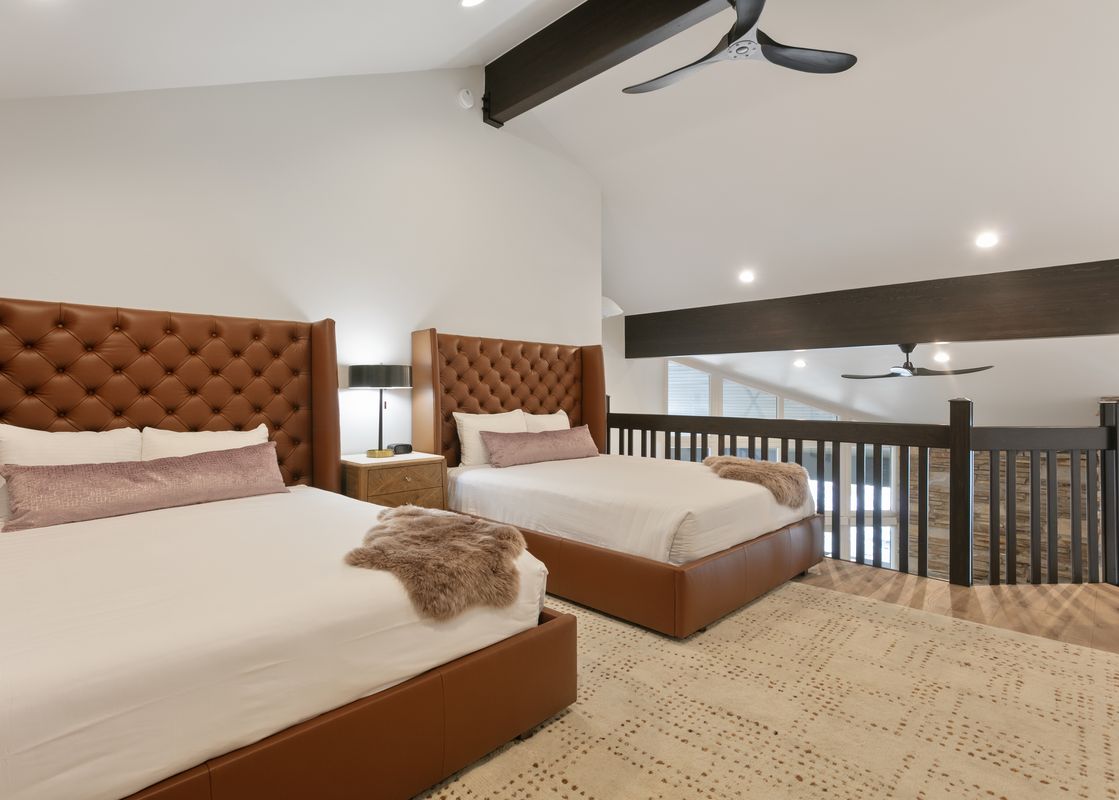 A bedroom in Powder Run B32 features two beds with brown leather tufted headboards, white bedding, and a textured rug, illuminated by recessed lighting and a ceiling fan.