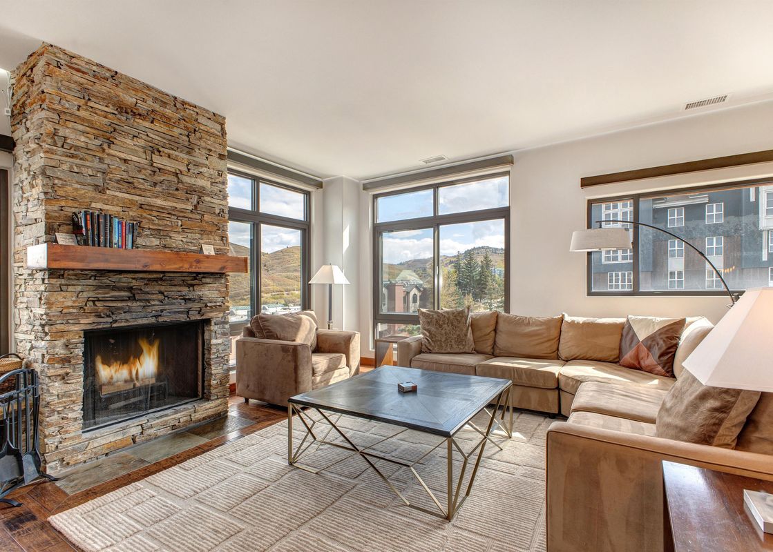 A spacious and elegantly furnished living room at Lowell 402B featuring a stone fireplace, a large L-shaped sofa, and armchairs, with expansive windows offering views of mountains and other buildings.