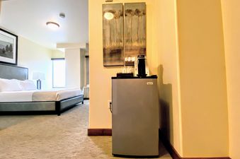 A cozy bedroom in Lowell 201A featuring a comfortable bed, bedside lamp, a mini-refrigerator with a coffee maker, and modern wall art, all bathed in soft light.