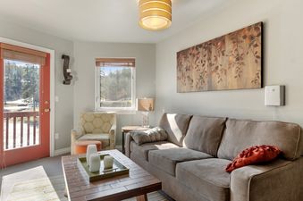 A cozy living room at Alpine Village 3 with a taupe sofa, patterned armchair, wooden coffee table, and an outdoor view through the door and window.