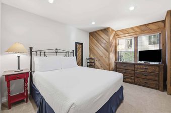A cozy bedroom with a king-sized bed, red nightstand, wooden dresser with a TV, and unique wooden wall paneling.