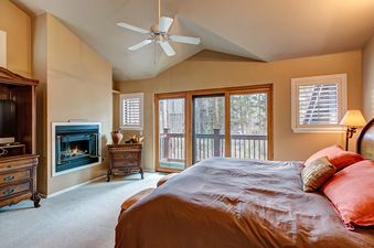 A bedroom with a king-size bed, a fireplace, and a view of the forest through a sliding glass door.