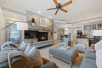 A cozy and modern living room at The Charter D1205 features a stone-faced fireplace with a television, comfortable seating, and an open-concept layout leading to a kitchen.