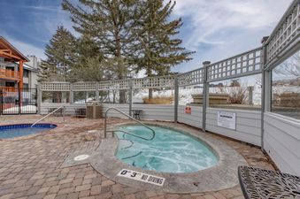Two outdoor hot tubs at Snow Flower 65 Park City are surrounded by a white lattice fence, evergreen trees, and a multi-story building, set against a cloudy blue sky.