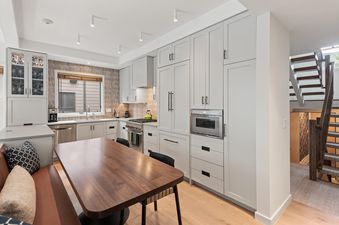A modern kitchen with light gray cabinets, a wooden dining table, and a built-in bench, with a staircase visible in the background.