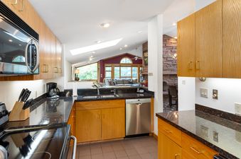 A modern kitchen with a black granite countertop and wooden cabinets includes a microwave, sink, and dishwasher, offering an open view into the living area with large windows.