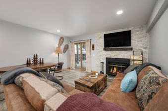 A cozy living area featuring a comfortable brown sofa with decorative pillows, a rustic wooden coffee table, and a glowing fireplace, with snowshoes decorating the wall.