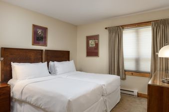 A cozy bedroom with two twin beds featuring white linens, wooden headboards, framed art, and a large window with drapes.